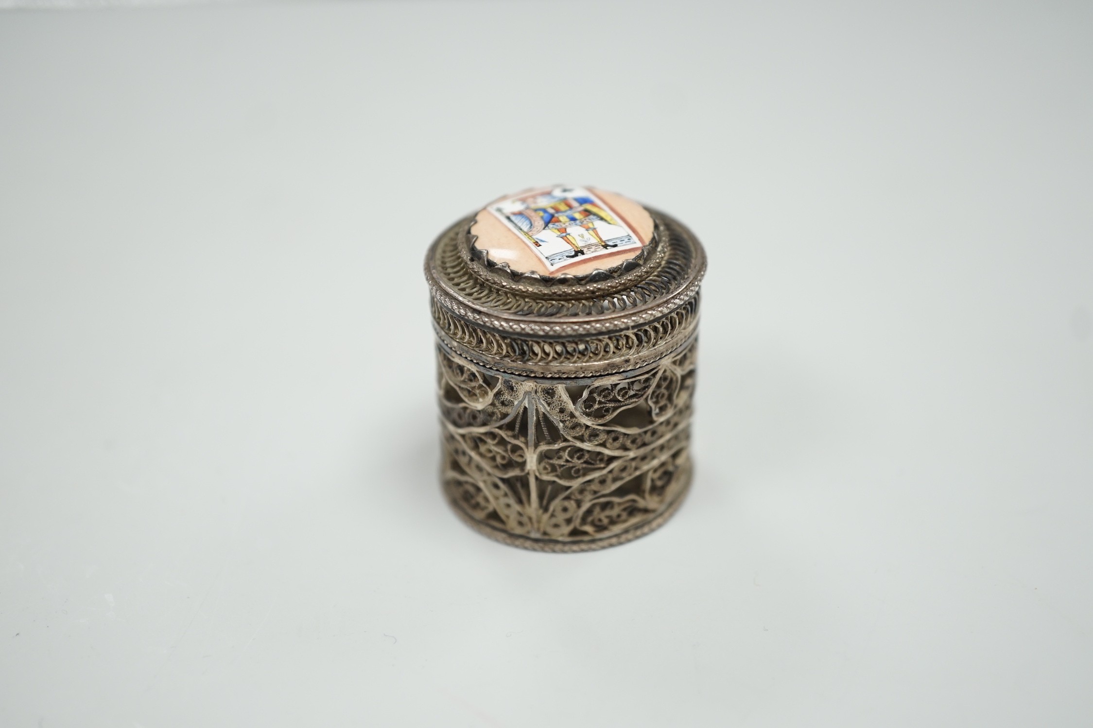 A continental filigree white metal and enamelled cylindrical pill box, the cover enamelled with a picture playing card, height 32mm.